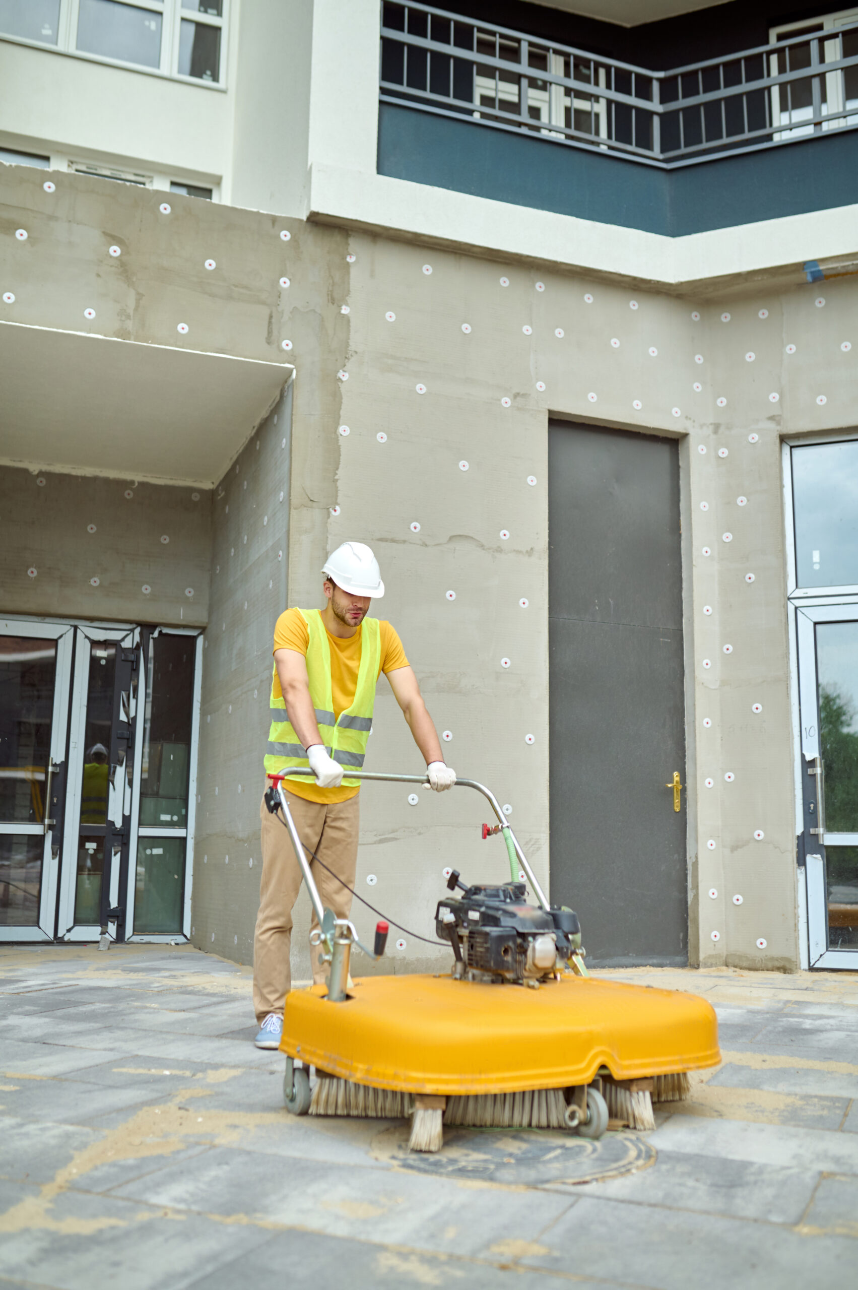Man operating special equipment cleaning construction site Cleaning. Man in protective helmet and gloves operating special equipment with brush cleaning construction site in new housing complex outdoors